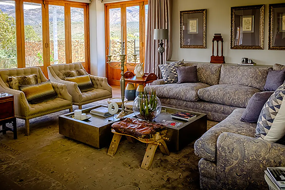 Perfect Hideaways property for sale: A lounge area with a large couch, and two olive-green leather chairs around a coffee table. The expansive farm is situated in the Karoo, and the surrounding countryside is visible through the lounge windows. Olive Farm in Prince Albert.