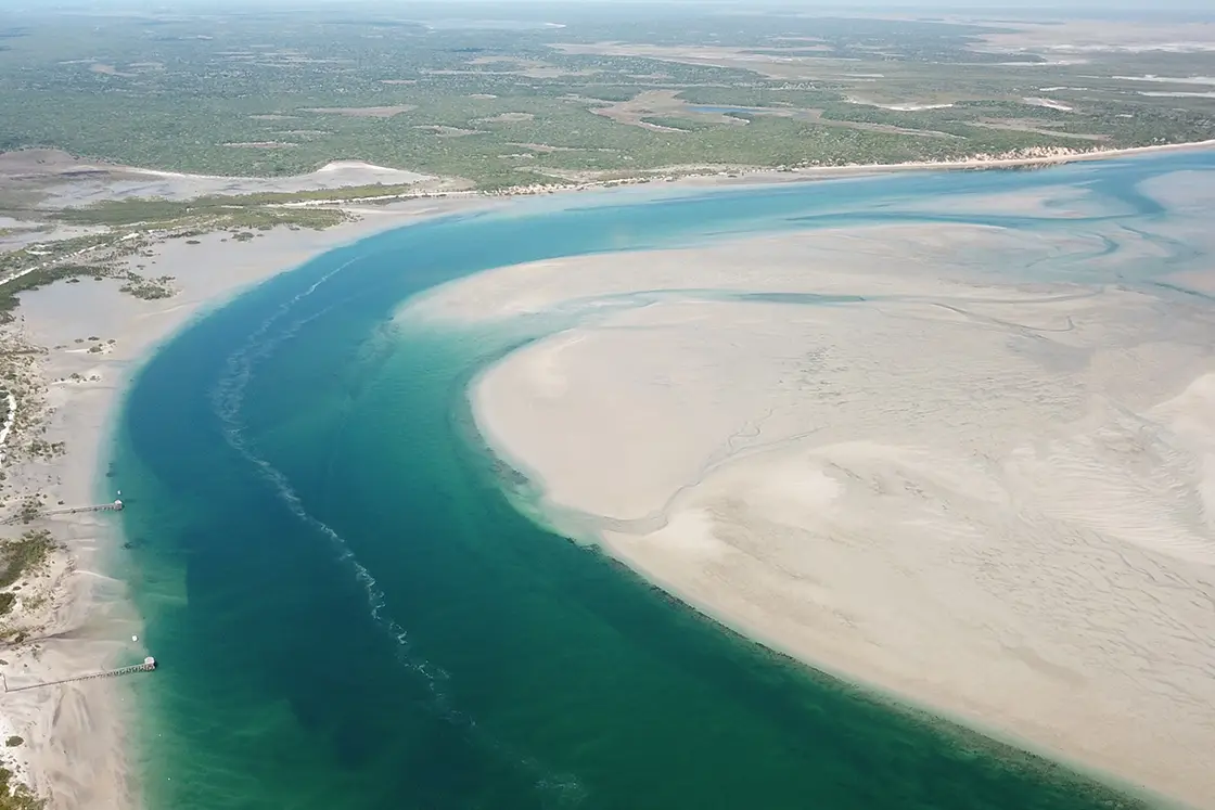 Perfect Hideaways real estate property: An aerial photograph of a waterway with coastal vegetation on one side of the waterway, and a shallow sand bank on the other. Two docks extend into the waterline. The Sanctuary Private Reserve, Mozambique.