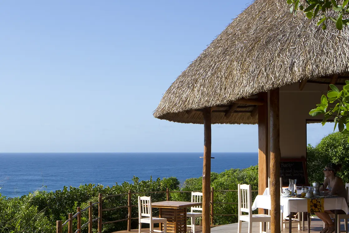 Perfect Hideaways real estate property: The dining area of the main lodge with wooden tables and chairs and a person sitting enjoying the view of the ocean. Dunes de Dovela Lodge, Mozambique.