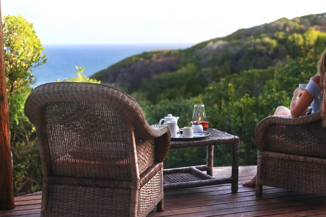 Perfect Hideaways real estate property: Two woven chairs with a person sitting in one, a coffee table in front with mugs, a teapot, and a lantern, with a sea view and hilltops in the distance. Dunes de Dovela Lodge, Mozambique.