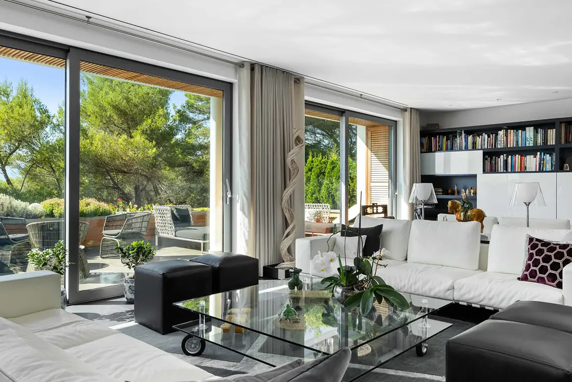 Perfect Hideaways real estate property: A lounge area with contemporary furnishings in black-and-white tones, featuring comfortable couches around a glass coffee table. Glass sliding doors provide access to the terrace, with outdoor seating and lush surrounding vegetation. Located at La Vue, in Mougins.