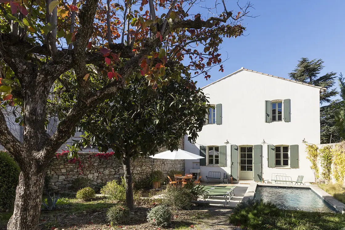 Perfect Hideaways property for sale: The French villa, as seen from the garden area with trees coloured in reds and greens, and an outdoor seating area beside the sparkling swimming pool. The villa itself features white exterior walls, and charming shutter windows with green shutters. France, Villa Sancti.