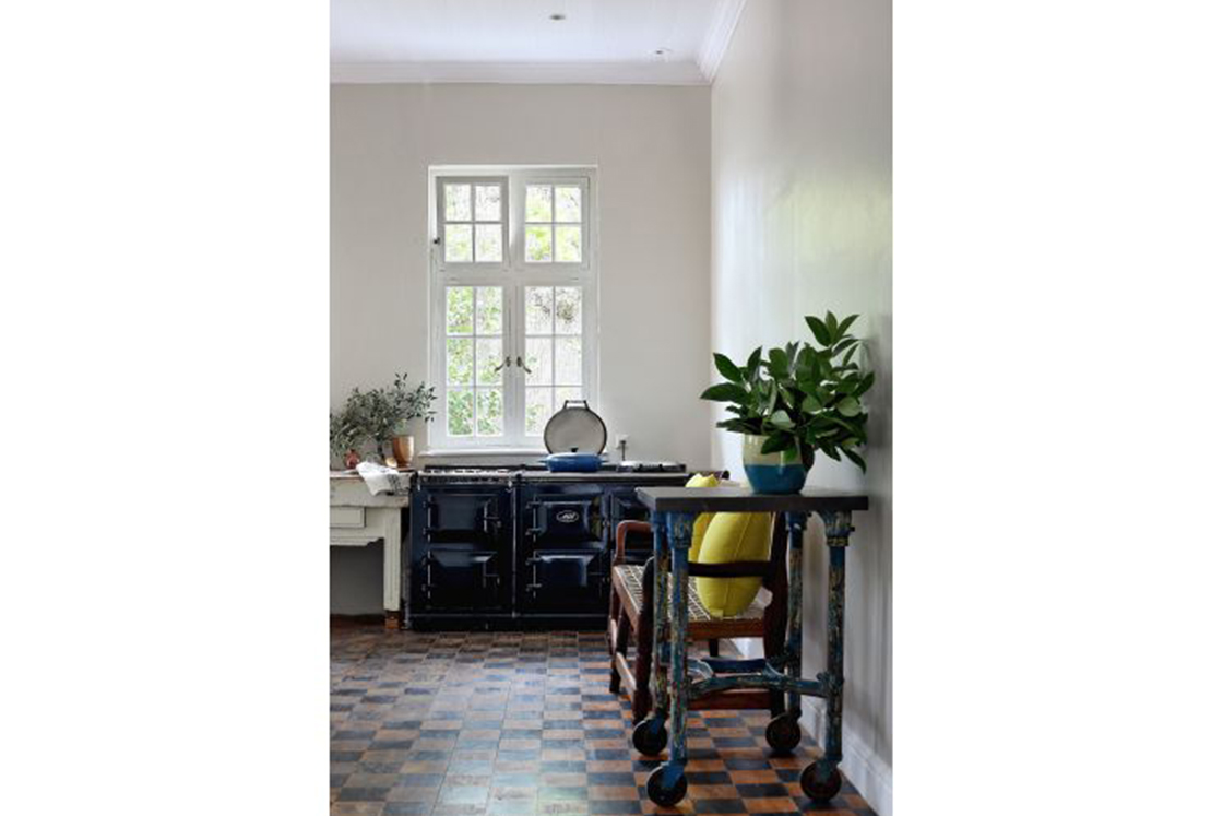 Sunlit kitchen area with rustic decor elements seen in a woven bench, portable table, and neutral table surface surrounding a 6-door Aga stove.