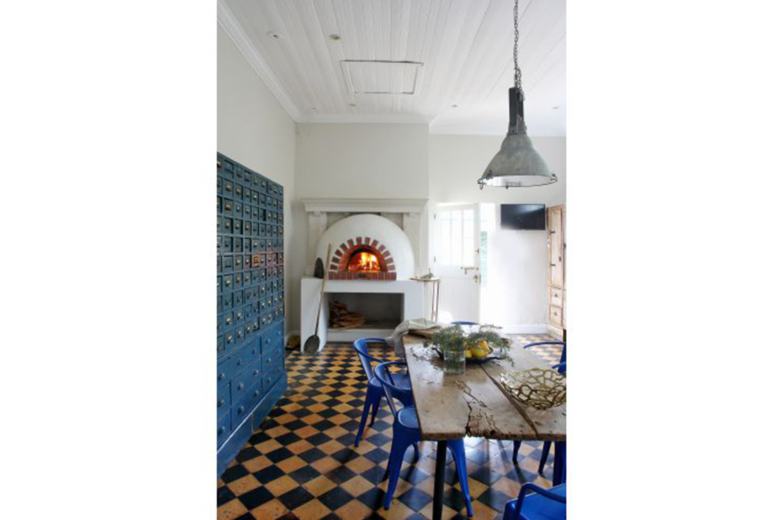 Kitchen with dining and preparation area featuring newly added pizza oven, aged checkerboard floor, rustic table with metal chairs and industrial storage unit echoing the heritage style of the home.