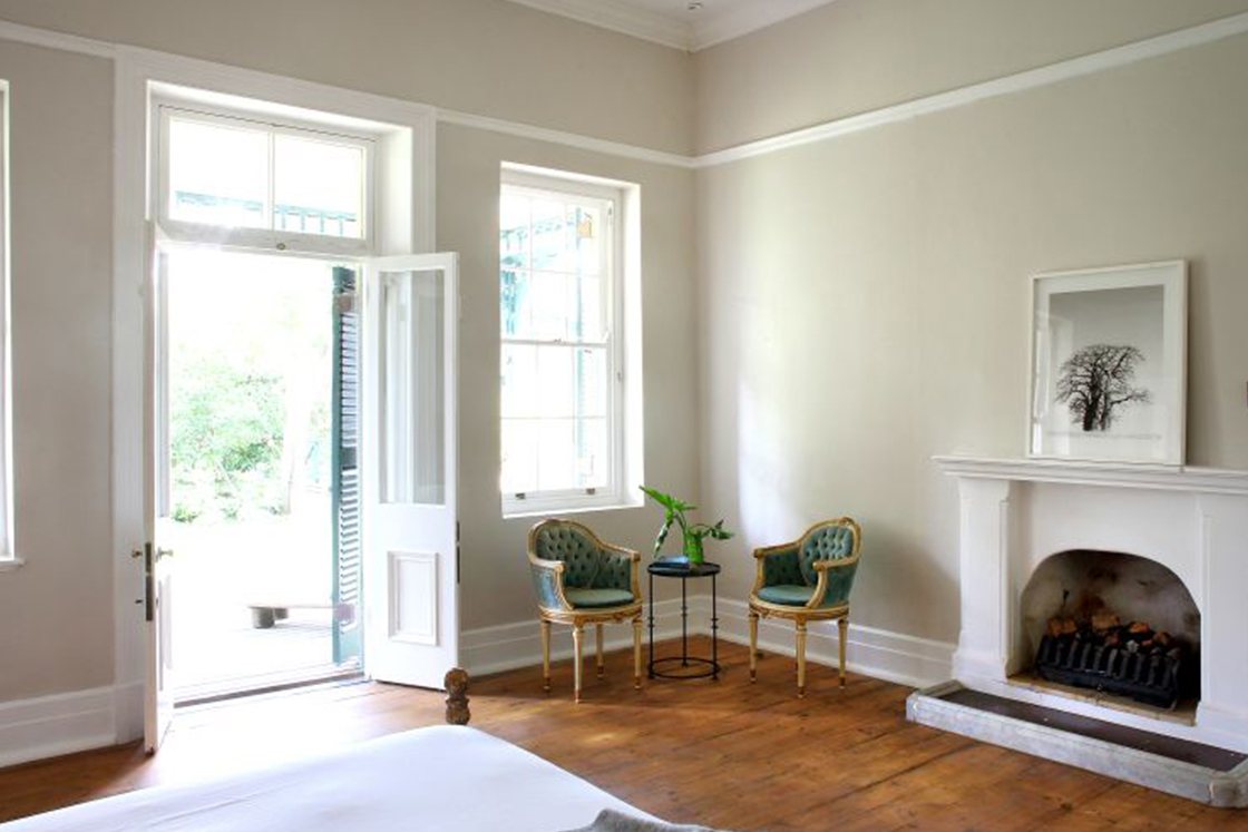 Light shining through elegant French doorway with direct garden access. Original Oregon pine floor and indoor fireplace provide a contrasting warmth to the neutral tones of the room, while antique chairs offer a comfortable seating area.