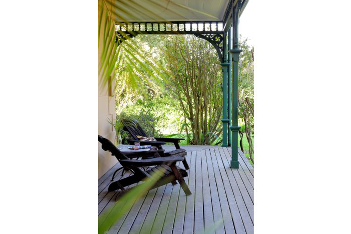 A wrap-around porch featuring two dark wood outdoor chairs facing out into the garden area. Wrought iron arches provide support for the bonnet roof.