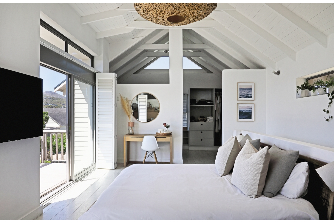 A modern bedroom that includes a large bed and a television, with white wooden beams, desk and walk-in cupboard.