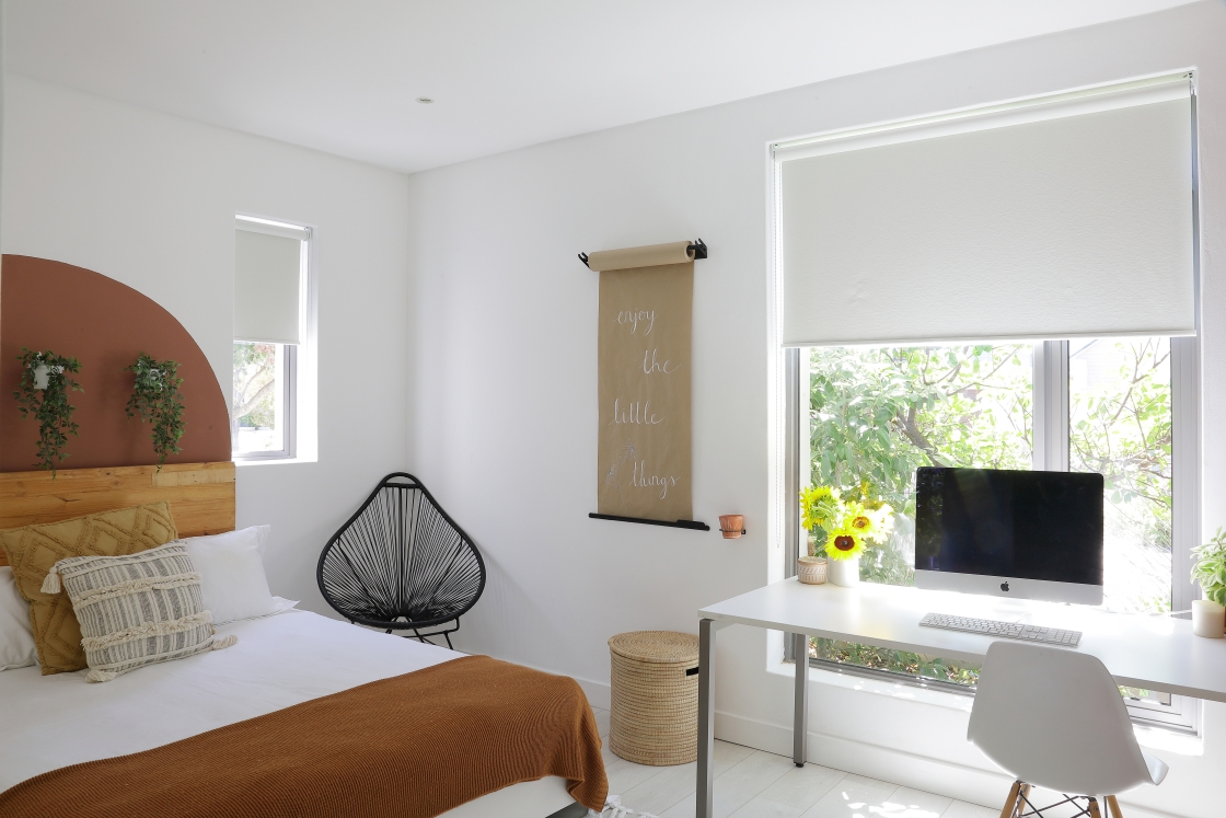 A bedroom scene displaying a bed with a terracotta headboard, a desk for work, and a window with a view of greenery.