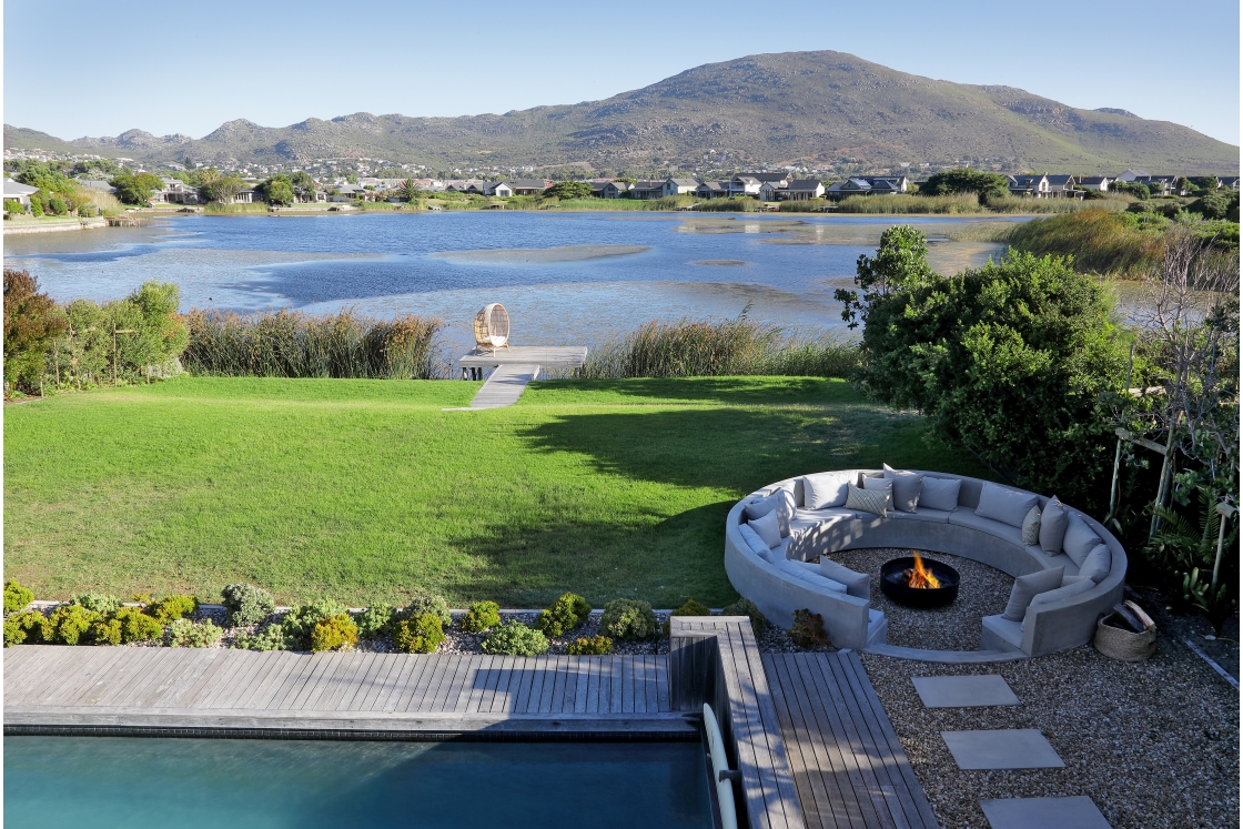 Aerial view showcasing a pool and lake, with a cozy fire pit area nearby, surrounded by lush greenery.