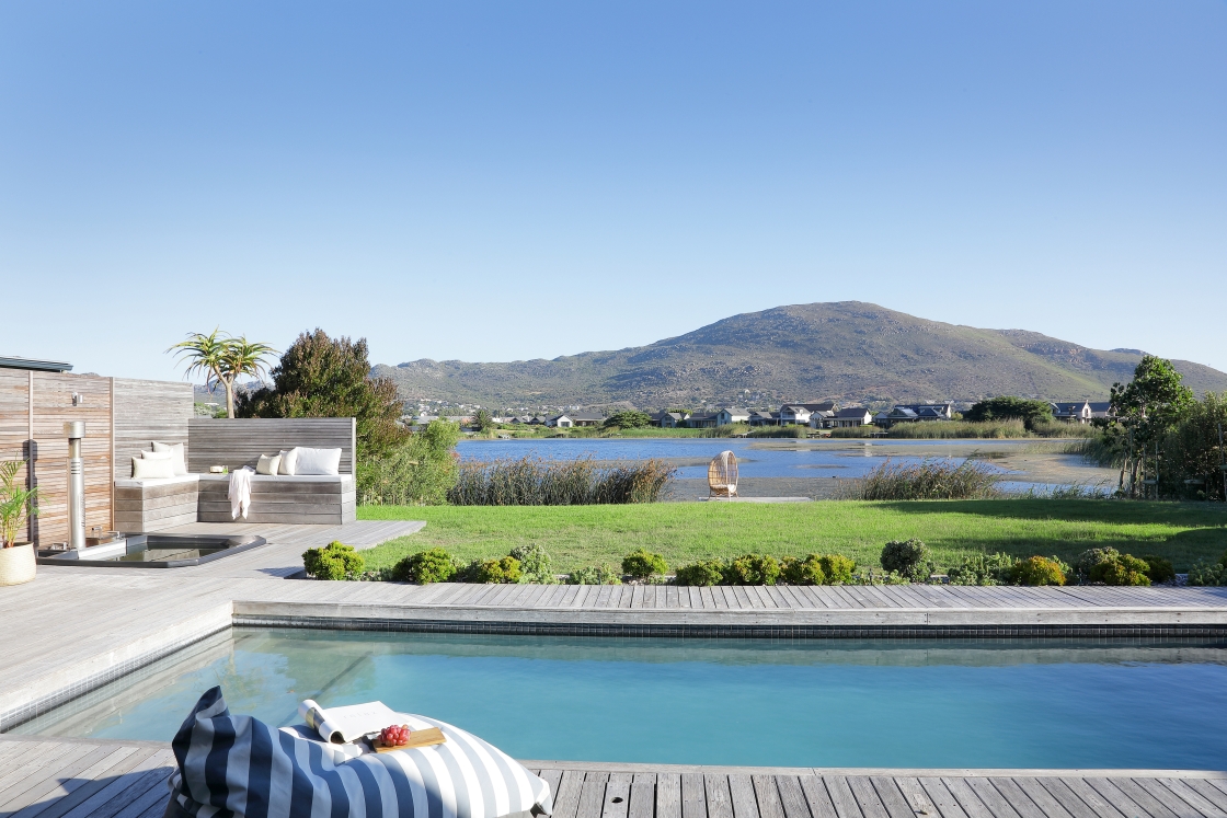 A pool with a view of a calm lake and mountains, with a wooden deck leading out onto green lawn.