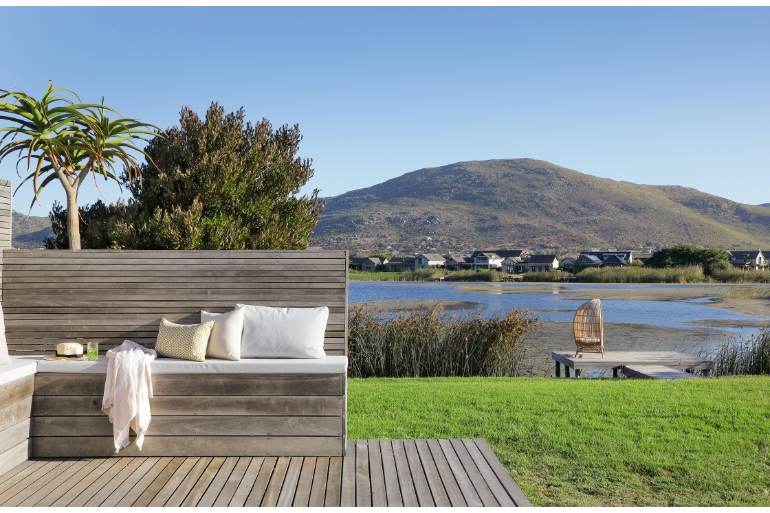 A wooden bench on a deck, offering a scenic view of a lake and mountains, with green lawn nearby.