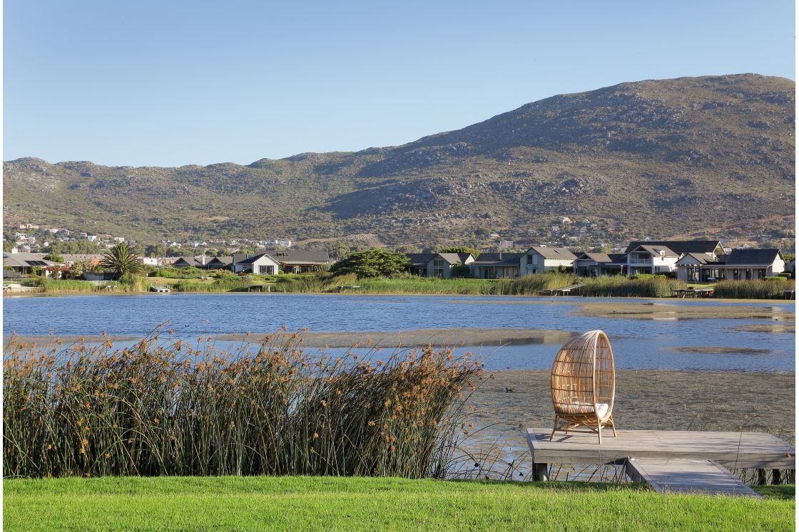 A body of water with a wooden jetty and wooden chair surrounded by lush greenery, houses on the estate and distant hills under a clear blue sky.