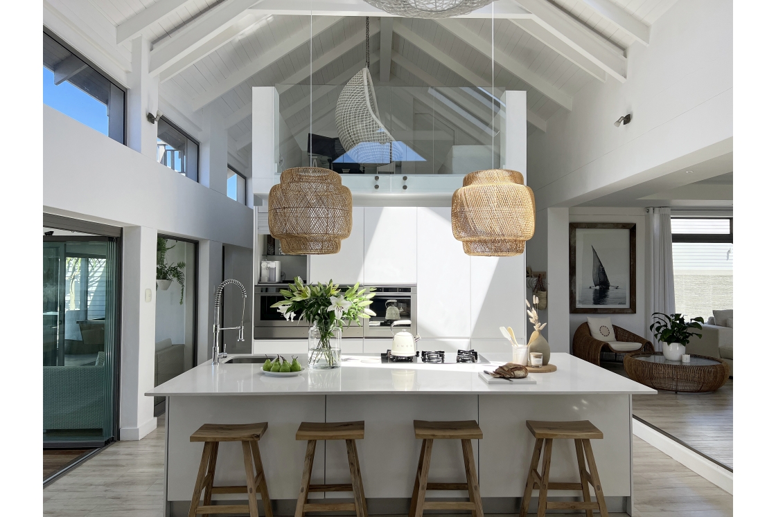 An open-plan kitchen with a white, marble island and wooden stools with high white wooden ceilings.