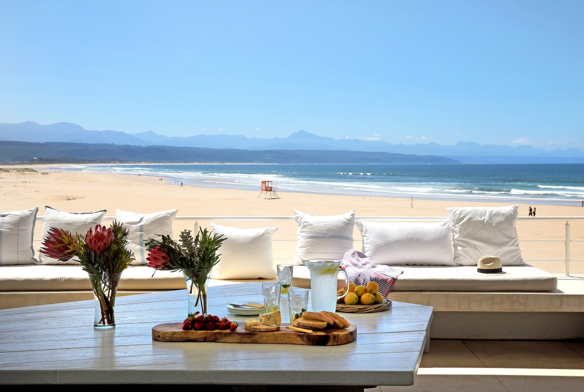 Perfect Hideaways real estate property: The terrace of a beach house, featuring a white geometric table with a floral arrangement of Proteas, a charcuterie board, and lemonade, overlooking Lookout Beach in Plettenberg Bay. Roga Rock, Plettenberg Bay.