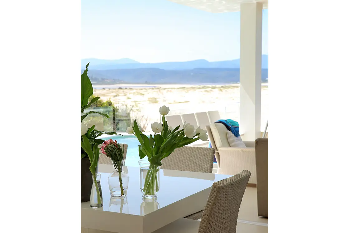 Perfect Hideaways real estate property: A close-up of the dining table, focusing on a flower arrangement. The dining area is adjacent to the pool and outdoor patio. Roga Rock, Plettenberg Bay.