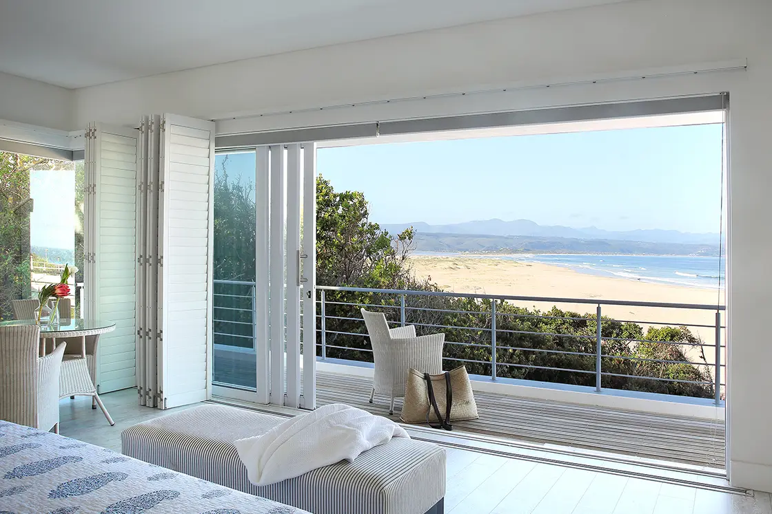 Perfect Hideaways real estate property: A bedroom area with shuttered sliding doors, a table and chairs, and an outdoor deck, featuring views of Lookout Beach, along the Garden Route. Roga Rock, Plettenberg Bay.