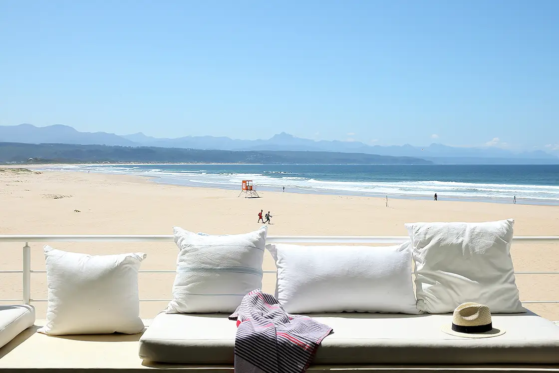 Perfect Hideaways real estate property: A balcony overlooking a long stretch of beach, with booth seating and a railing. The coastline of The Garden Route in the background. Roga Rock, Plettenberg Bay.