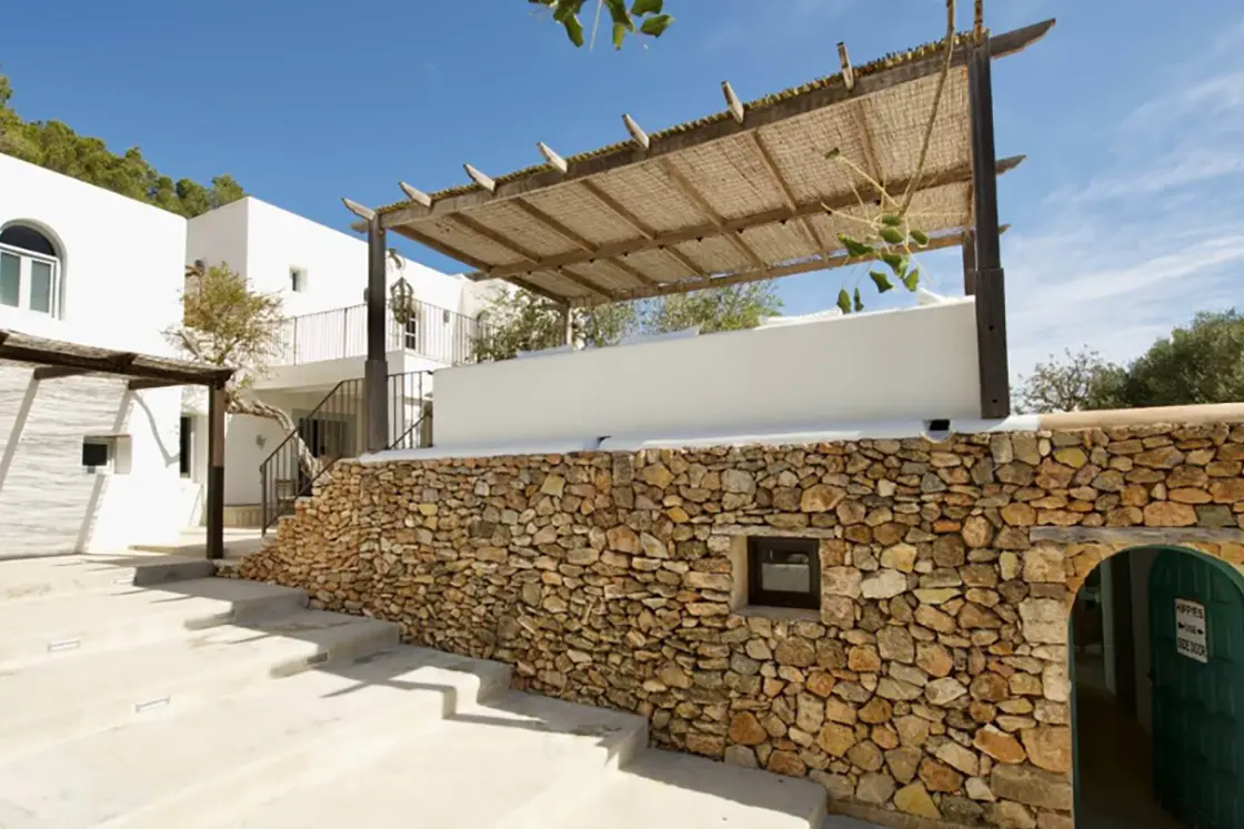 Perfect Hideaways real estate property: An entranceway to the Finca home, which features large steps beside a natural stone wall, a green arched door into the lower floors, and multiple pergolas which offer shaded seating areas for guests. Ibiza, Ibizan Finca.