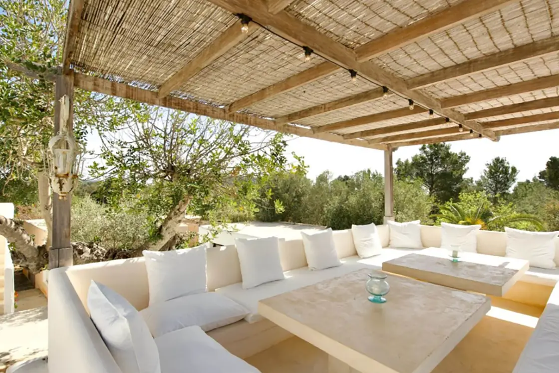 Perfect Hideaways real estate property: An outdoor seating area with booth seating and wraparound couches, shaded by a pergola, and surrounded by lush vegetation. Ibiza, Ibizan Finca.