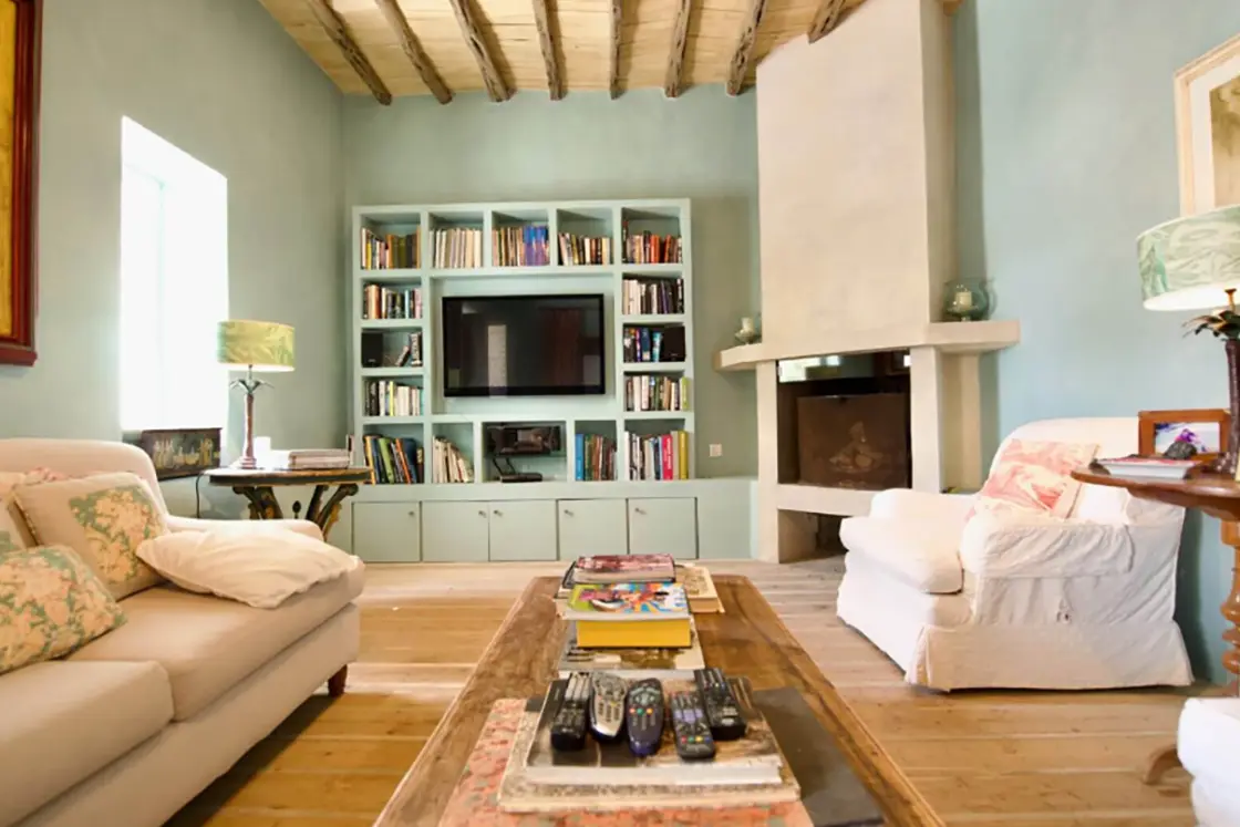 Perfect Hideaways real estate property: A living area of the Spanish home, featuring muted blue tones in the wall colour, as well as wooden floor and ceilings. Comfortable couches, an indoor fireplace, and a bookshelf. Ibiza, Ibizan Finca.