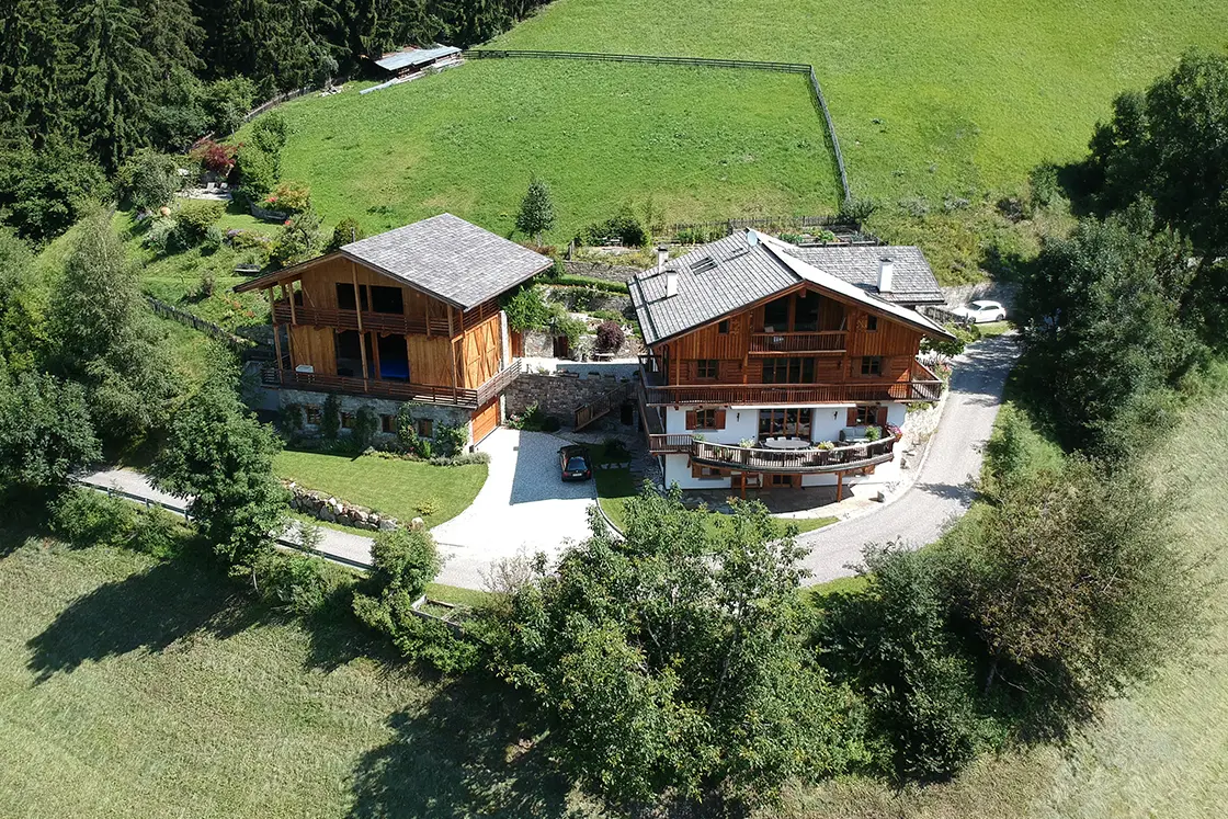 Perfect Hideaways real estate property: An aerial view of a pair of Italian chalets, featuring a private garden, a large grass area above the chalets, and a forest surrounding the property. Ragginerhof, Italy.