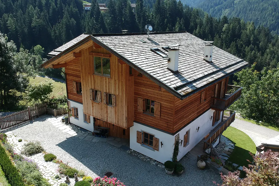 Perfect Hideaways real estate property: A four-story Italian chalet, featuring a mainly wooden exterior, angled tile roof, and lush vegetation in the background. Ragginerhof, Italy.