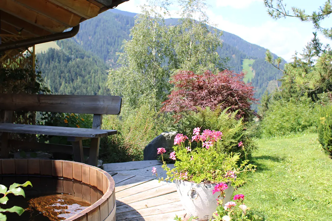 Perfect Hideaways real estate property: A four-person Canadian hot tub outside of the Italian chalet, featuring decorative flowers and vegetation in the garden, and views of the Dolomites in the background. Ragginerhof, Italy.