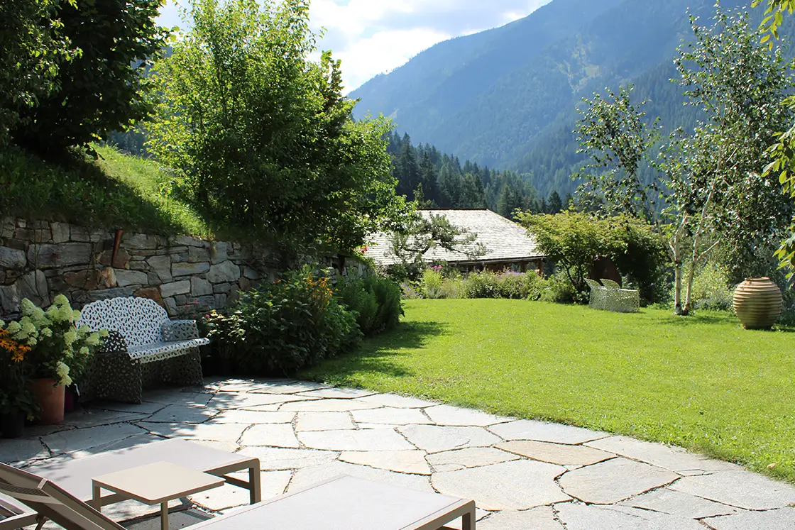 Perfect Hideaways real estate property: An outdoor terrace with a natural stone floor and a lawn leading towards the second chalet, in the background, views of the Italian countryside. Ragginerhof, Italy.