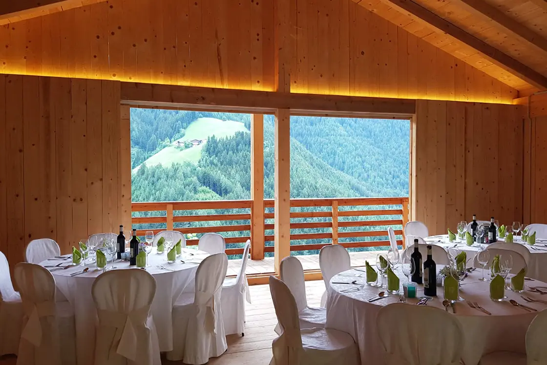 Perfect Hideaways real estate property: A dining room area, featuring three circular tables set to entertain multiple guests, while the chalet-style room is constructed out of natural wood, and has a balcony with views of the Dolomite mountain range. Ragginerhof, Italy.