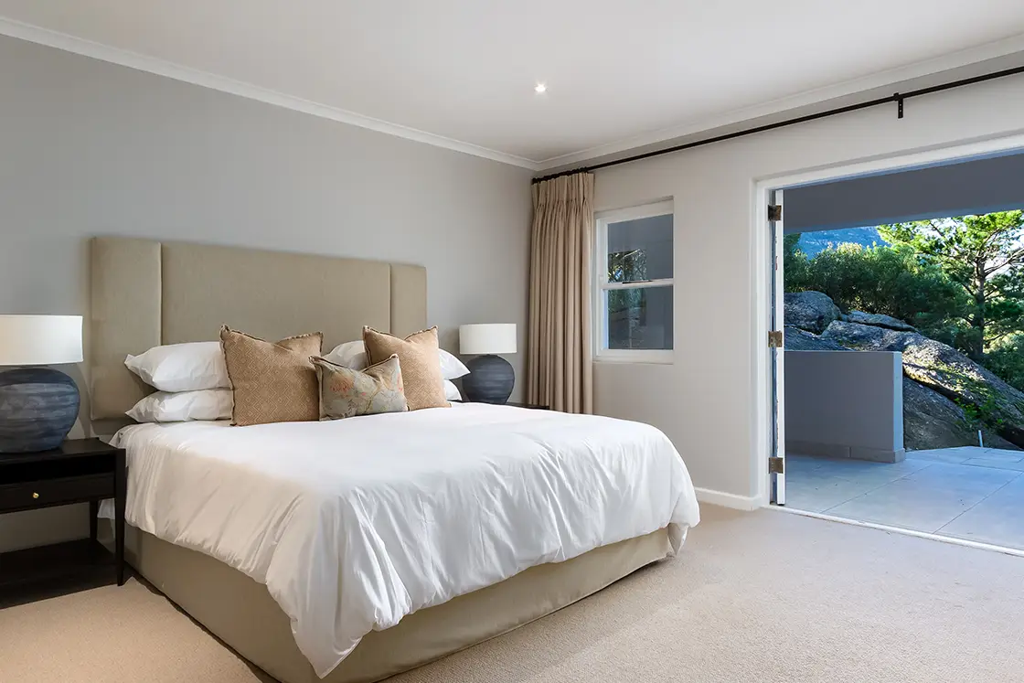 Perfect Hideaways real estate property: A minimally furnished bedroom with a double bed, featuring dark-wood side tables with ceramic lamps. Open double doors lead to a covered patio. The Berg Manor and Villas, Hout Bay.