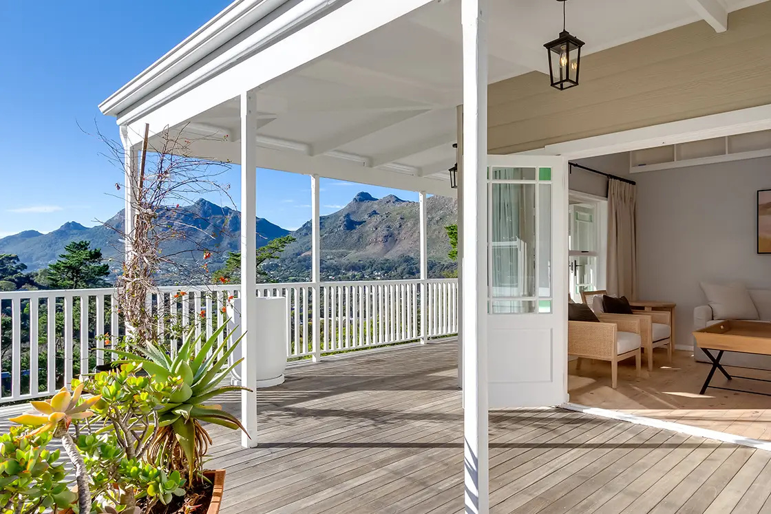 Perfect Hideaways real estate property: A lounge area with open double doors leading onto a wooden deck, showcasing views of mountains in the background. The Berg Manor and Villas, Hout Bay.