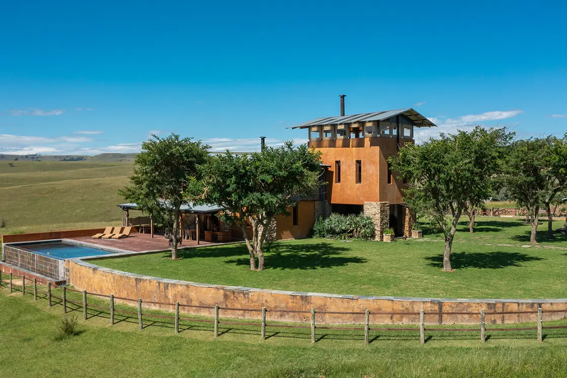 Perfect Hideaways real estate property: A guest lodge with orange walls and natural stone supports, featuring an outdoor pool, a manicured lawn with trees, and rolling hills in the background. Zulu Waters Game Reserve, Natal Midlands.