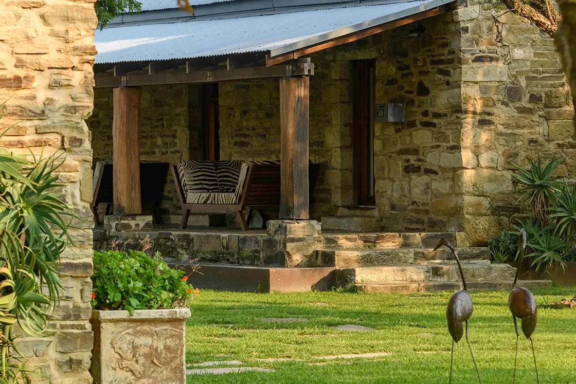 Perfect Hideaways real estate property: A small guest cottage with natural stone exterior walls and a covered patio, featuring zebra-patterned chairs and a manicured lawn with two metal bird statues. Zulu Waters Game Reserve, Natal Midlands.