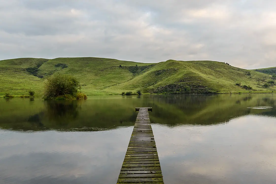 Perfect Hideaways real estate property: A moss-covered jetty extends into a still lake, with the green hills across the lake reflected in the water. Zulu Waters Game Reserve, Natal Midlands.