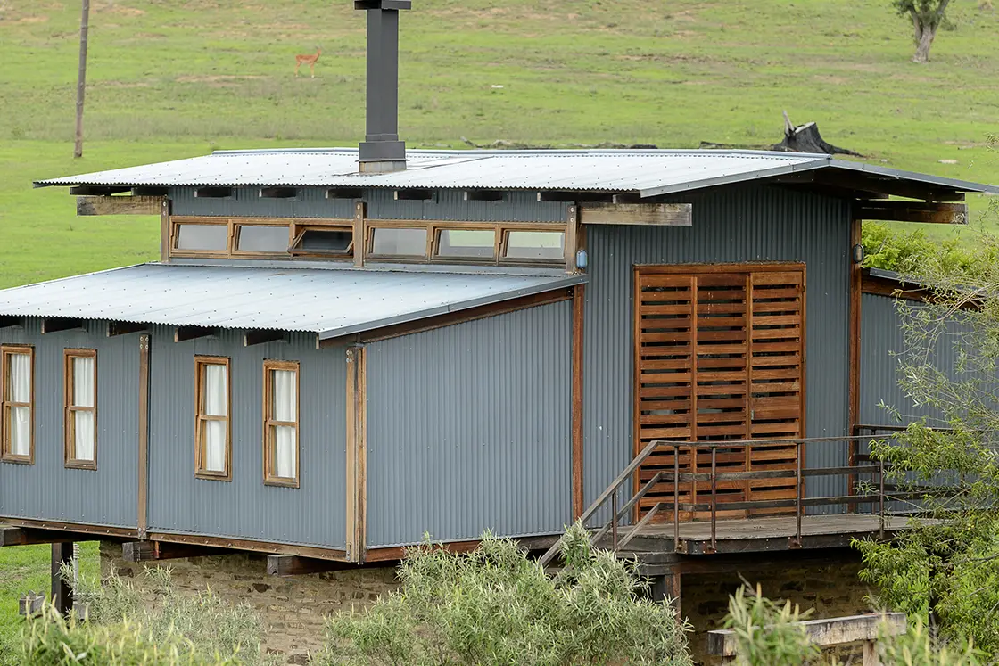 Perfect Hideaways real estate property: A corrugated metal lodge with a sliding wooden doorway, wooden window frames, and a chimney, featuring views of the surrounding grassland. Zulu Waters Game Reserve, Natal Midlands.