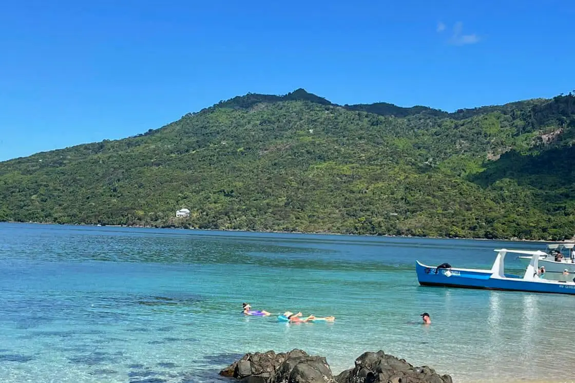 Perfect Hideaways real estate property: A beach in Madagascar, with swimmers in the shallows, a nearby water taxi, and a large hill with dense vegetation in the background. Komba Eco Lodge, Madagascar.