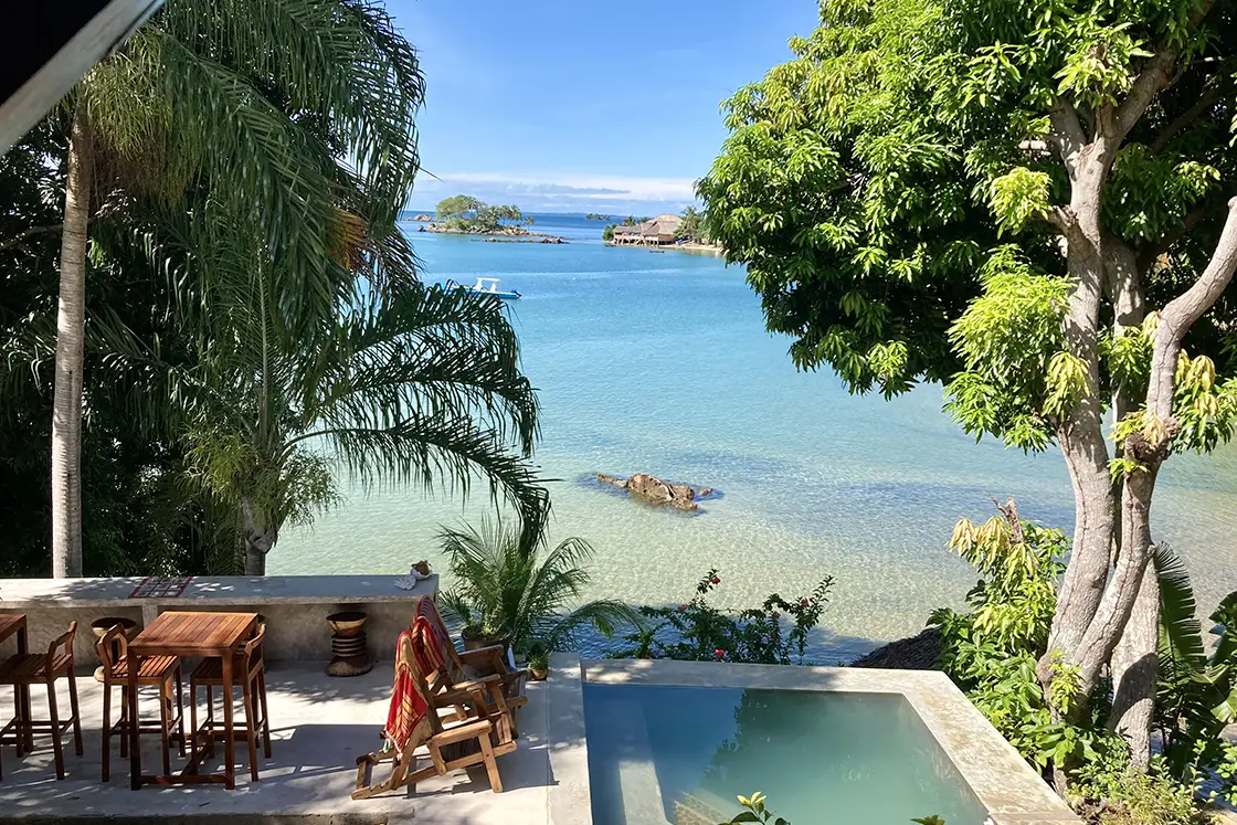 Perfect Hideaways real estate property: An outdoor dining area and a pool surrounded by wooden chairs, featuring views of the clear blue water and small tropical islands in the background. Komba Eco Lodge, Madagascar.