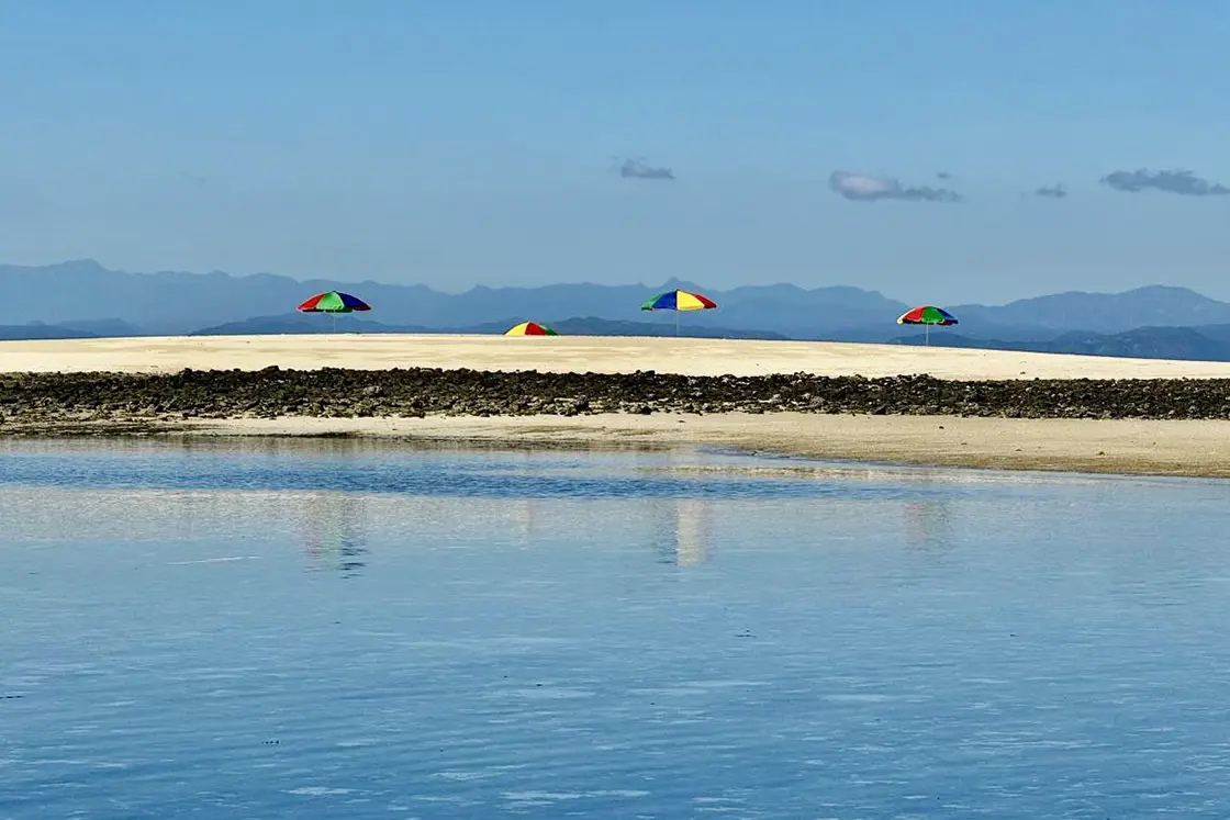 Perfect Hideaways real estate property: Clear shallow water with a beach and umbrellas, featuring views of mountains in the background. Komba Eco Lodge, Madagascar.