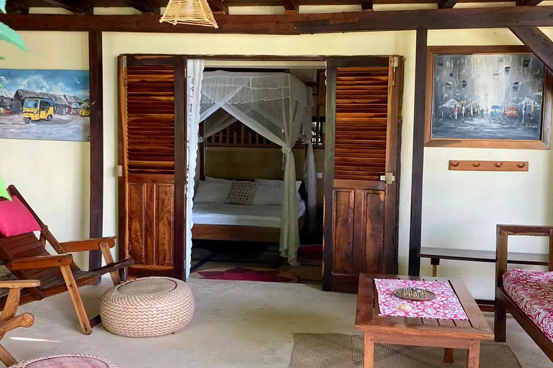 Perfect Hideaways real estate property: A living area, featuring wood furniture, hand-made decor, and art pieces decorating the walls, a wooden doorway leads to a bedroom with a four-poster bed. Komba Eco Lodge, Madagascar.