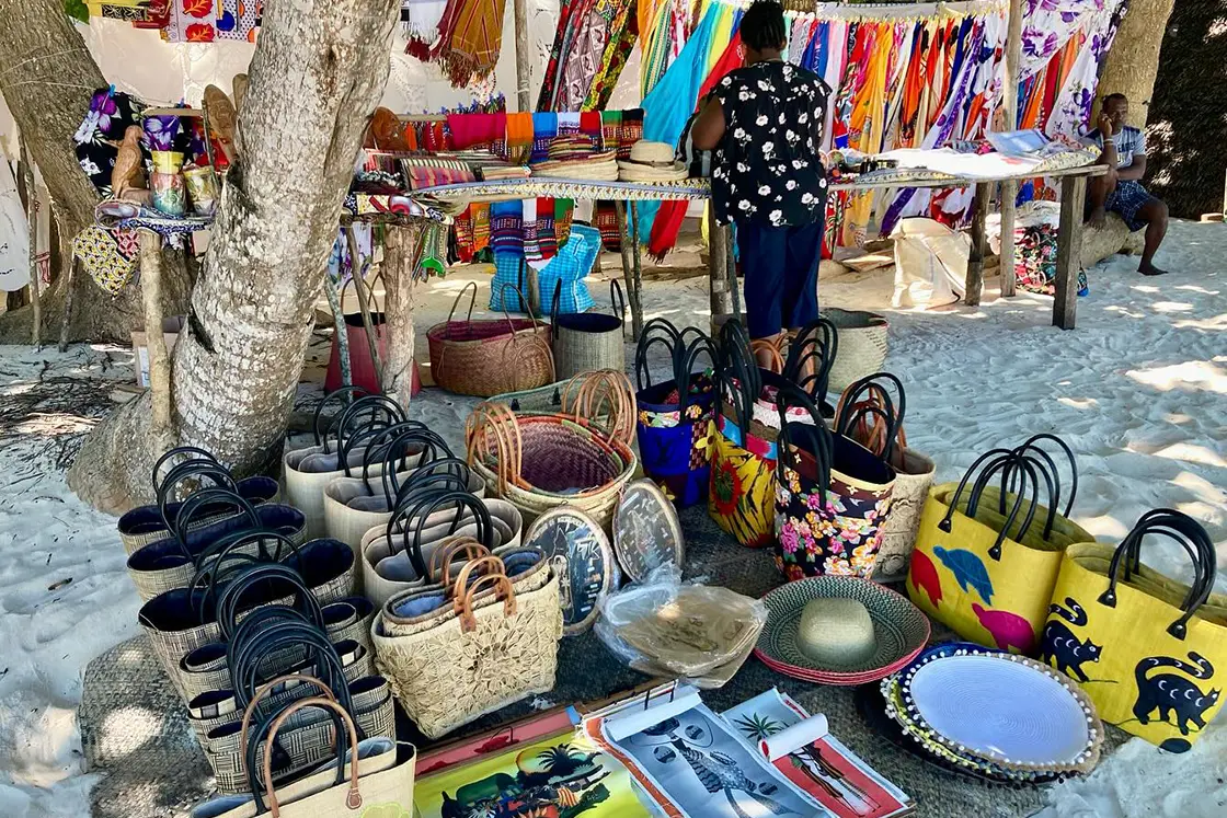 Perfect Hideaways real estate property: A local woman on a beach surrounded by tropical trees, showcasing the handmade goods for sale. Komba Eco Lodge, Madagascar.