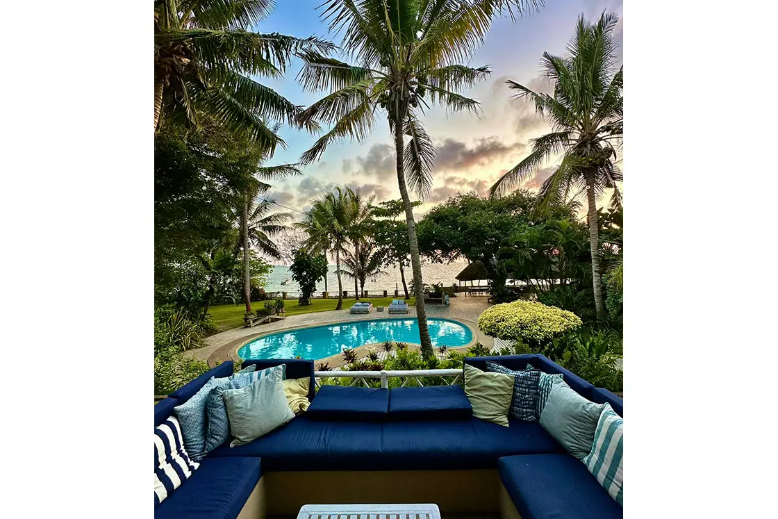 Perfect Hideaways real estate property: A navy blue conversation couch overlooking the outdoor patio of the guest lodge, which includes a pool, seating, gazebos, lush vegetation, and a deep-water channel in the background. Villa 1b Beach House, Vilanculos.