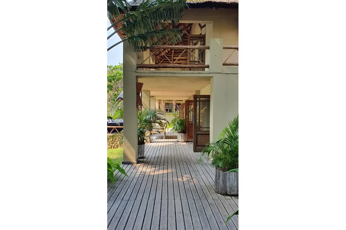 Perfect Hideaways real estate property: A two-story guest lodge, with thatch-roofing, featuring an outdoor deck, and potted local vegetation, as well as surrounding palm trees. Villa 1b Beach House, Vilanculos.