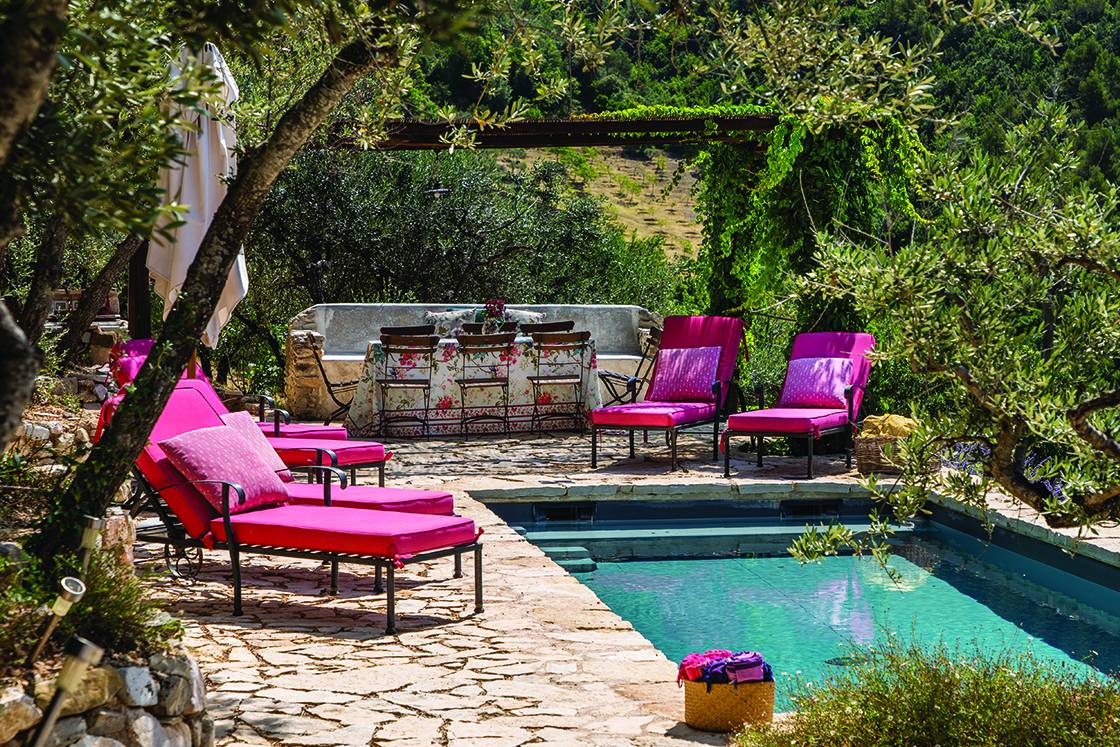 Perfect Hideaways real estate property: A sunbathing area next to a sparkling swimming pool, which features natural stone tiles and sun chairs, behind the pool is an outdoor dining area sheltered by a vine-covered pergola. Casa Di Led in Spoleto.