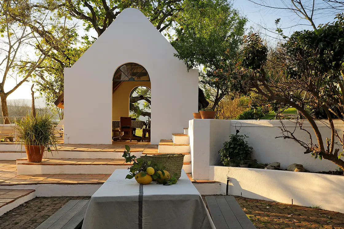 Perfect Hideaways real estate property: A courtyard table adorned with lemons, linen tablecloth, and woven basket. A farm style 'Summer House' features in the background. Zeekoegat Farm in Riversdale.