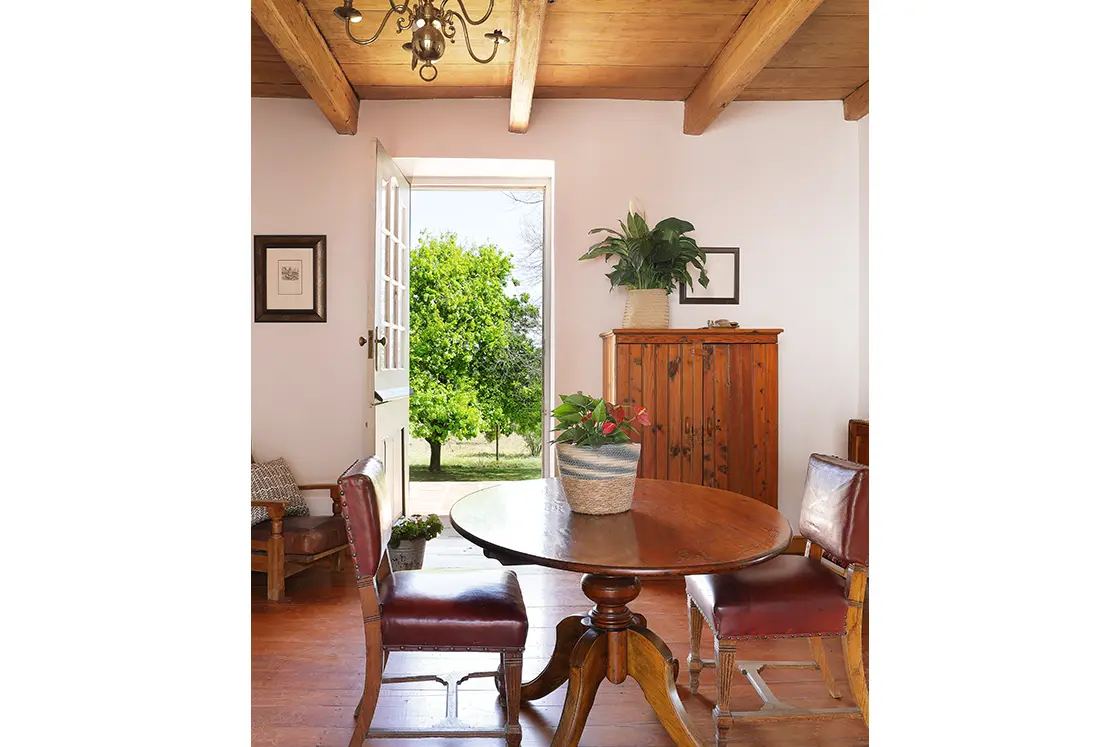 Perfect Hideaways real estate property: Interior view of farmhouse, two leather chairs set around an ornate dining table, wooden cabinet against back wall, with garden view through back door. Zeekoegat Farm in Riversdale.