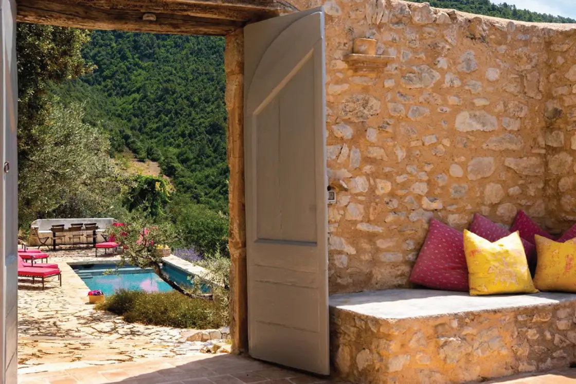 Perfect Hideaways real estate property: The natural stone exterior wall of an outdoor courtyard, with seating and an open double doorway. Pool and sun loungers with views of the lush hillside. Casa Di Led in Spoleto.