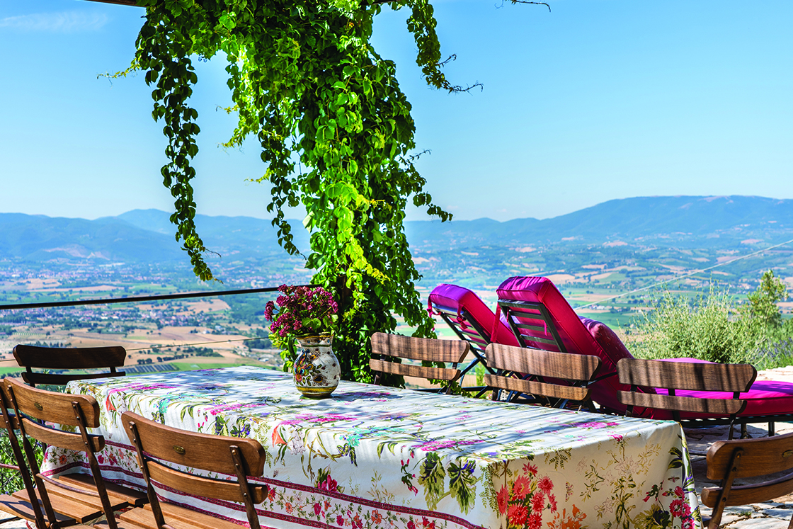 Perfect Hideaways real estate property: A pergola with green vines provides shade for the outdoor dining area. Floral table setting and dining chairs with views of the Italian countryside. Casa Di Led in Spoleto.