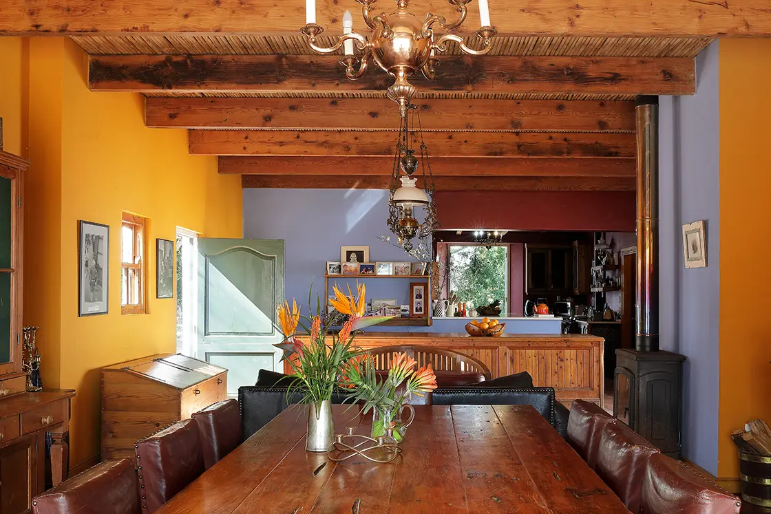 Perfect Hideaways real estate property: Large wooden table set with local flowers and leather chairs. Muted blue tones and orange walls complement the space. Zeekoegat Farm in Riversdale.