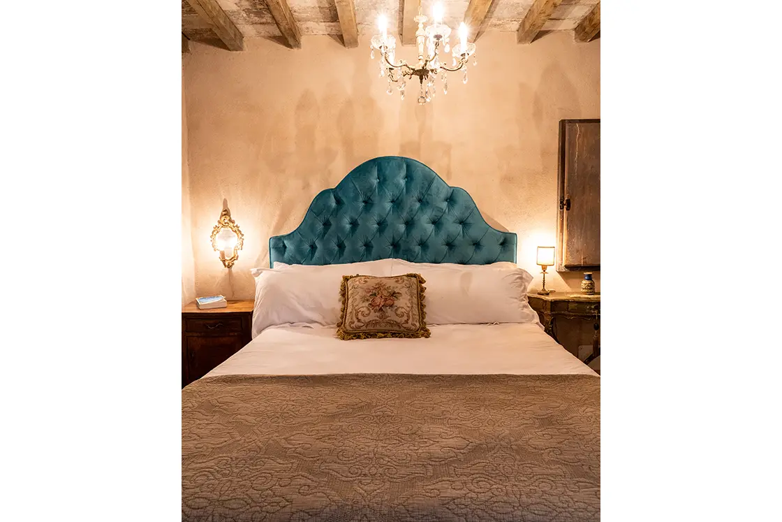 Perfect Hideaways real estate property: A double bed with a blue buttoned headboard and a decorative pillow. Ornate chandelier and bedside lighting. Casa Di Led in Spoleto.