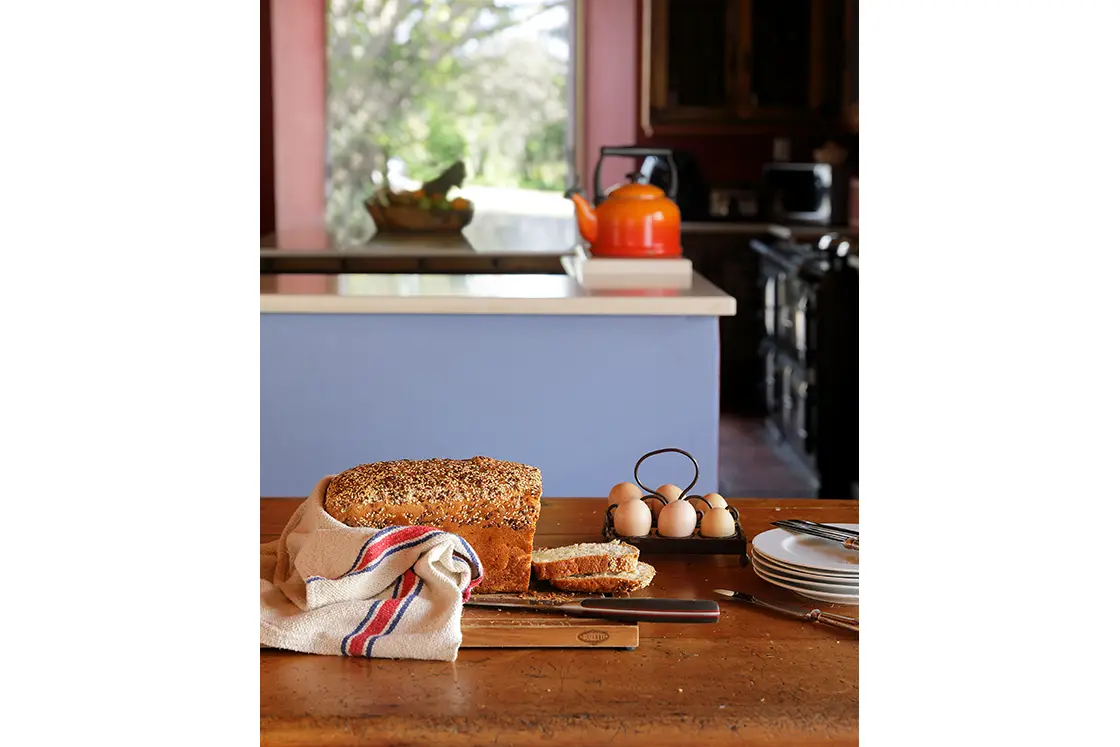 Perfect Hideaways real estate property: Farm style bread, eggs, and cutlery on a wooden table in the foreground. Blue kitchen island with kettle and garden view in the background. Zeekoegat Farm in Riversdale.